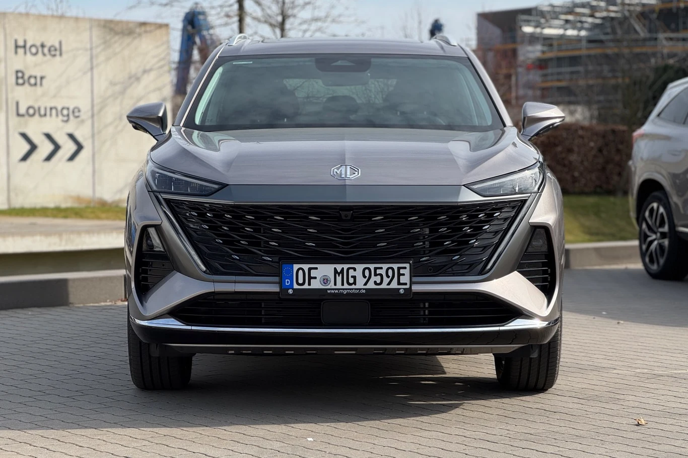 MG S9 będzie wyróżniać się na trasie przez swój dyskusyjny design nowoczesny srebrny SUV MG z wyrazistym, szerokim grillem i smukłymi reflektorami na tle miejskiego otoczenia, z widoczną tablicą rejestracyjną