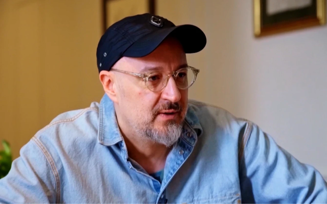 mężczyzna w jasnej dżinsowej koszuli i czarnej czapce z daszkiem, z krótką brodą i okularami, siedzący w pomieszczeniu o jasnych ścianach, w tle wiszące obrazy