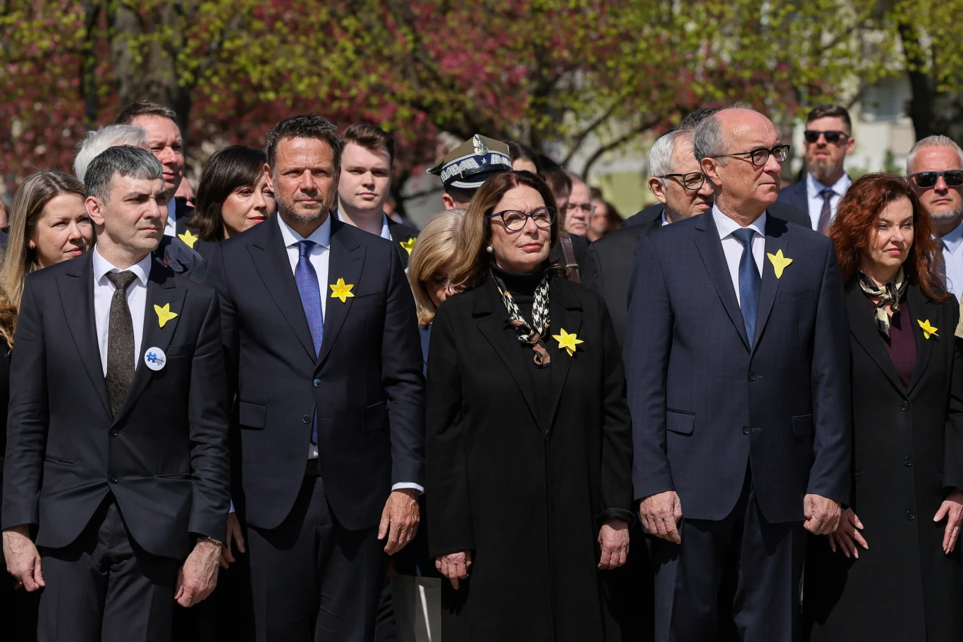 Grupa osób ubranych w eleganckie, ciemne ubrania stoi w skupieniu, większość z nich ma przypięte papierowe żółte żonkile, co wskazuje na uczestnictwo w upamiętnieniu o historycznym charakterze.