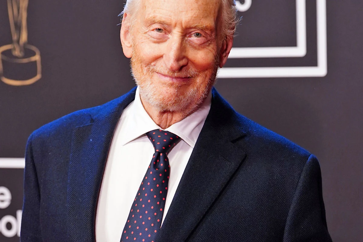 Charles Dance Charles Dance w granatowym garniturze, białej koszuli i czerwonym krawacie na tle czarnej ścianki z napisem.