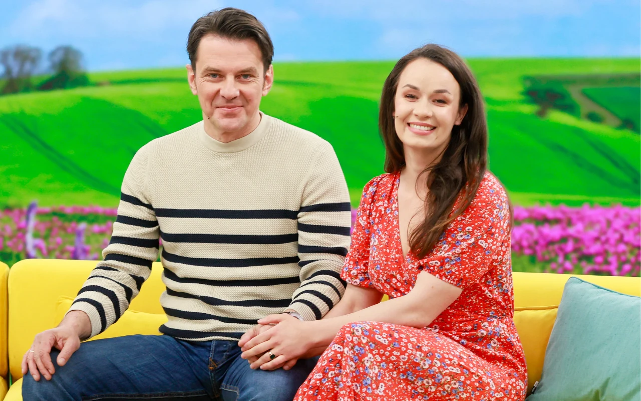 Dwie osoby siedzące razem na żółtej kanapie w kolorowym studio, mężczyzna w swetrze w paski trzyma za rękę uśmiechniętą kobietę w czerwonej sukience, w tle wyraźna grafika soczyście zielonych pól i kwitnących kwiatów.