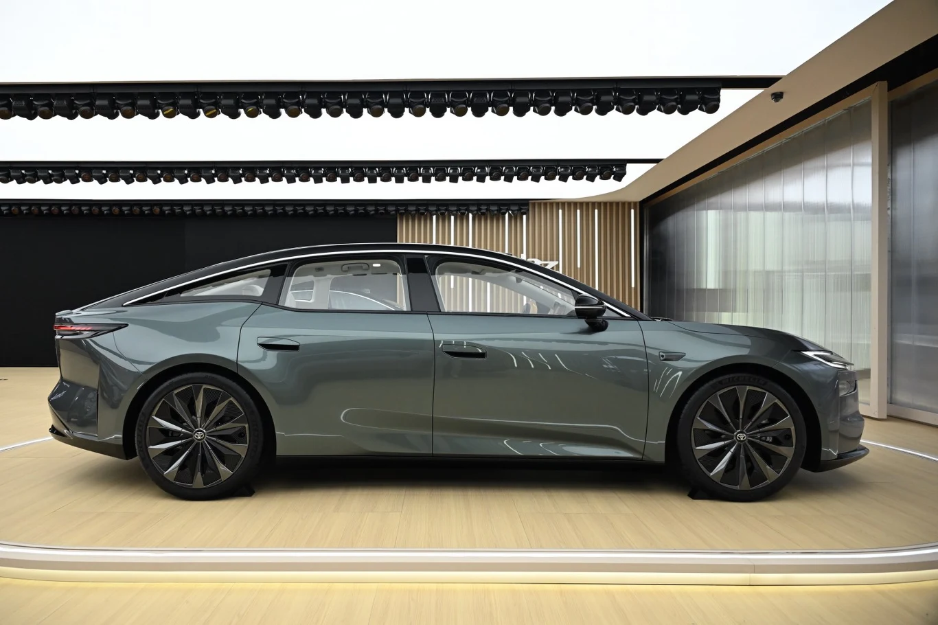 nowoczesny, luksusowy sedan elektryczny o opływowej sylwetce w kolorze ciemnej zieleni, z dużymi felgami aluminiowymi, osadzony w jasnym, minimalistycznym wnętrzu wystawowym