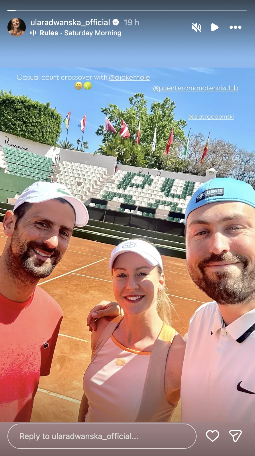 Urszula Radwańska spotkała na korcie Novaka Djokovicia Trójka osób w sportowym stroju uśmiecha się do wspólnego selfie na korcie tenisowym, w tle widoczne puste trybuny oraz zielona roślinność.