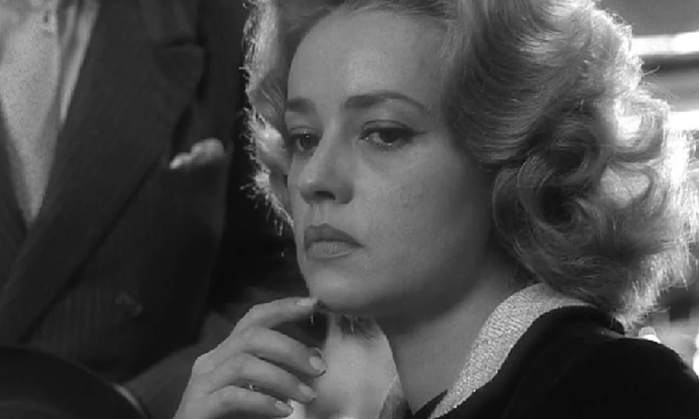 Kadr z filmu "Windą na szafot", fot. Timeless Film Festival Warsaw kobieta o jasnych, falowanych włosach z zamyśloną miną, w eleganckim ubraniu, opiera brodę na dłoni, w tle fragment sylwetki mężczyzny w garniturze