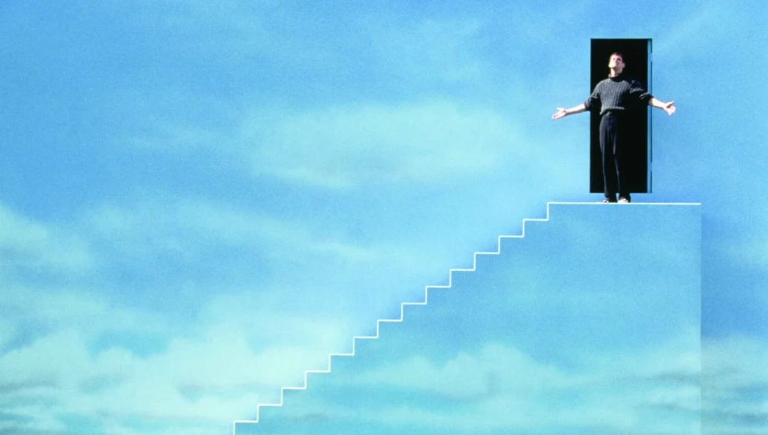 Kadr z filmu "Truman Show", fot. Timeless Film Festival Warsaw Kadr z filmu 'Truman Show'. Mężczyzna stoi w otwartych drzwiach na szczycie białych schodów, w tle niebo.