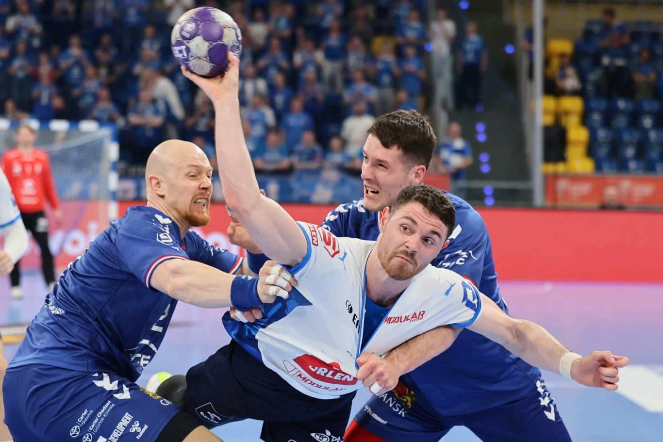 dynamiczna akcja piłki ręcznej, trzech zawodników walczących o piłkę, zawodnik w białej koszulce stara się rzucić piłkę, podczas gdy dwóch przeciwników w niebieskich strojach próbuje go powstrzymać