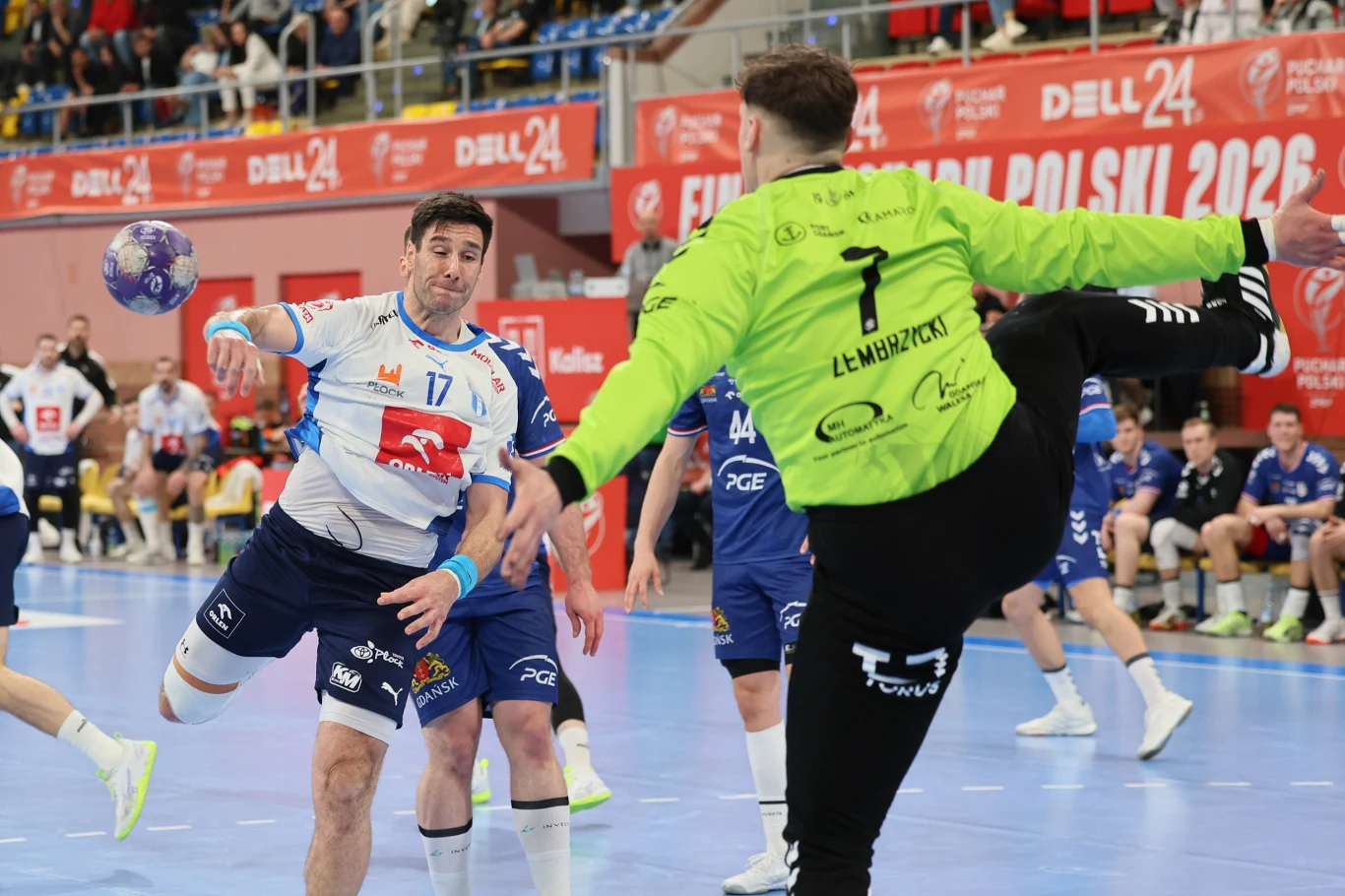 zawodnik w białej koszulce z numerem 17 rzuca piłkę ręczną w stronę bramkarza w jaskrawozielonym stroju, który dynamicznie próbuje obronić rzut podczas meczu piłki ręcznej na hali sportowej