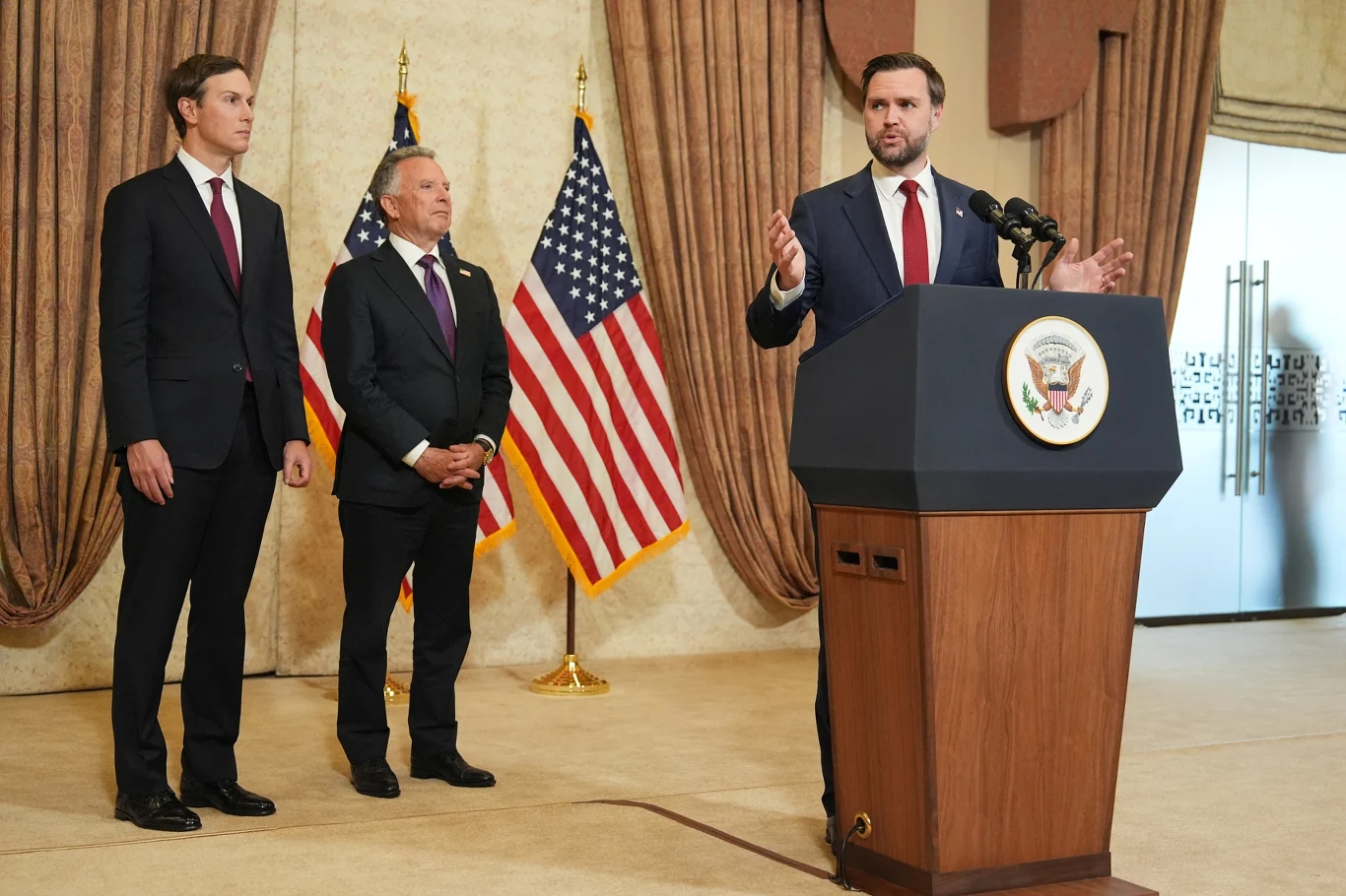 Negocjacje USA - Iran. Niejasności wokół obecności wiceprezydenta J.D. Vance'a Trzech mężczyzn w garniturach, jeden przemawia przy mównicy z pieczęcią USA, w tle dwie flagi amerykańskie