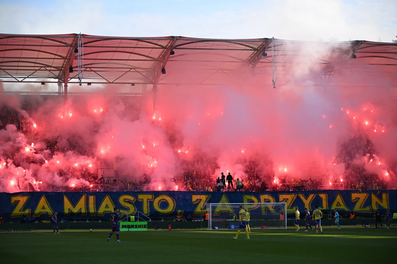 Kibice Arki Gdynia kibice na trybunach stadionu odpalają race, tworząc intensywną czerwoną poświatę i gęsty dym nad sektorami, w tle widoczny duży transparent z napisem, na pierwszym planie piłkarze na murawie oraz bramka