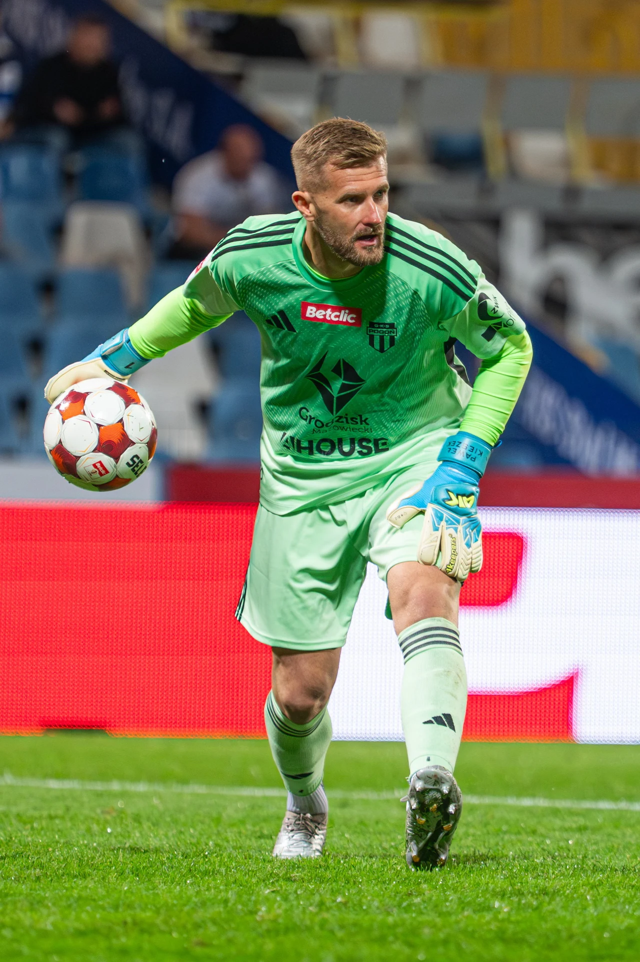 bramkarz w jasnozielonym stroju z piłką w lewej ręce przygotowuje się do wykopu na boisku piłkarskim podczas meczu, w tle zamazane trybuny ze światłami stadionowymi