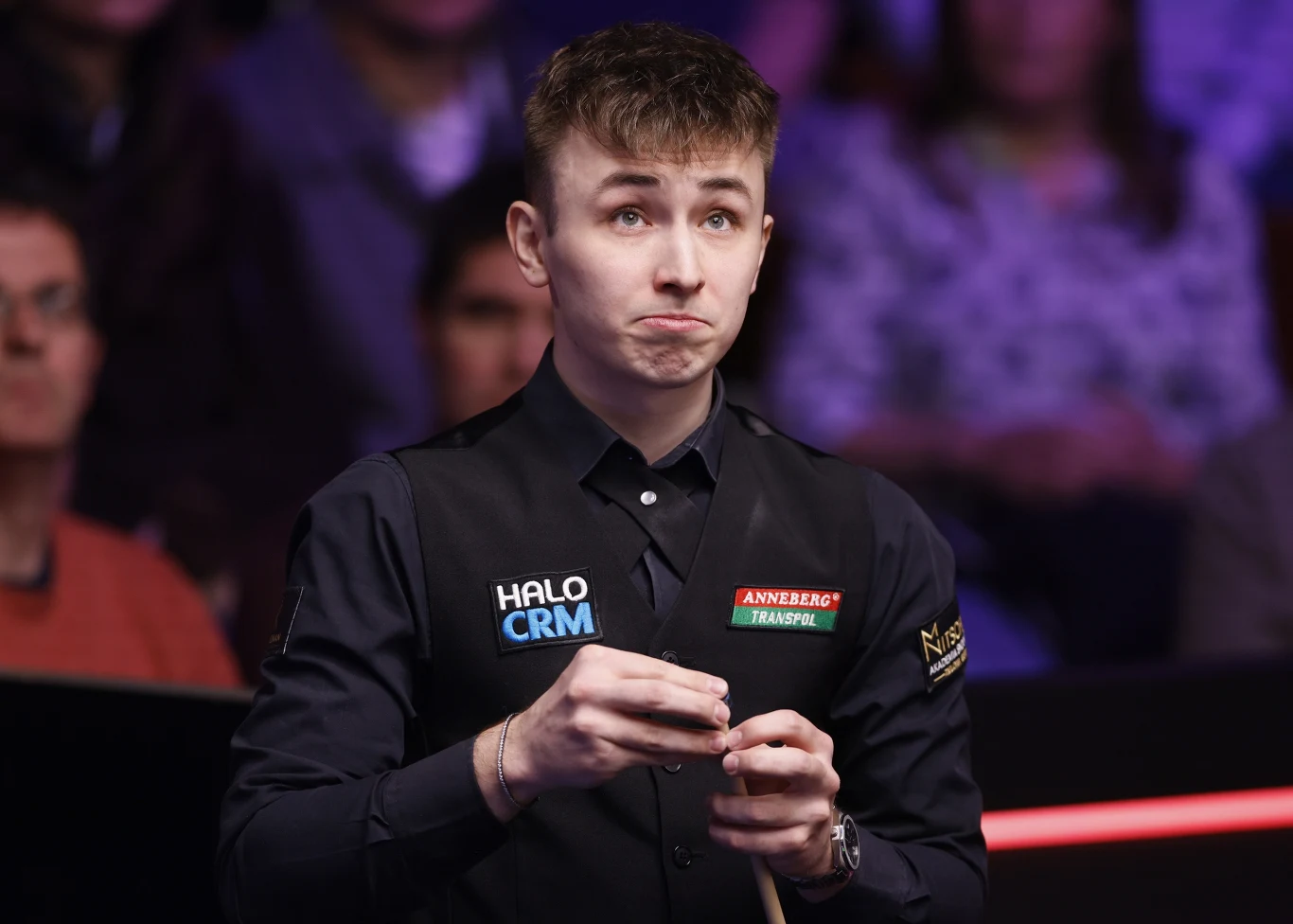 młody mężczyzna w ciemnej kamizelce z naszywkami sponsorskimi stoi przy stole snookerowym, trzymając kij i kredę, z poważnym wyrazem twarzy na tle zamazanej publiczności