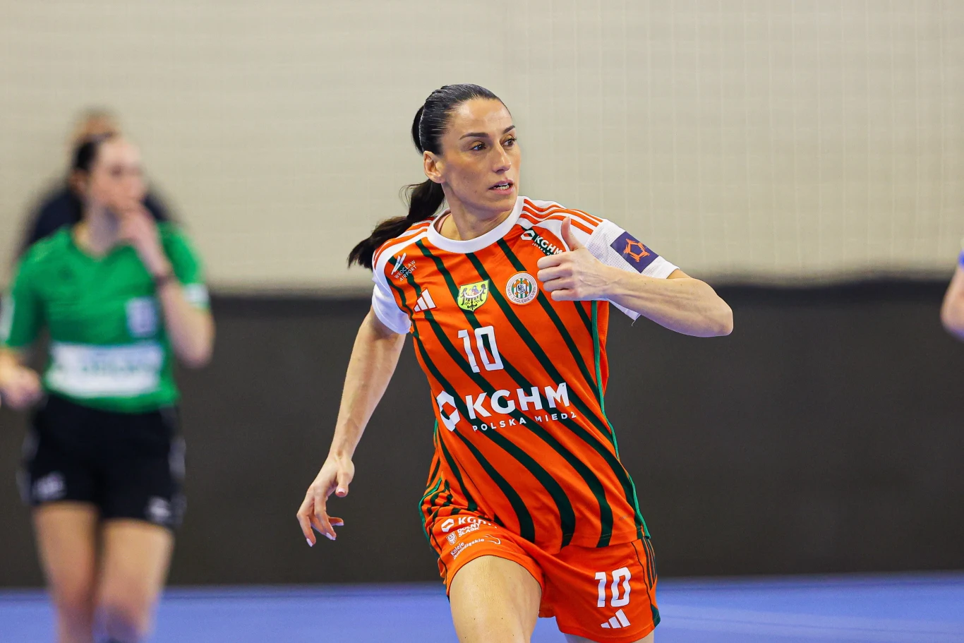 zawodniczka w pomarańczowo-czarnej koszulce z numerem 10 i opaską kapitana biegnąca po hali sportowej podczas meczu piłki ręcznej, w tle sędzia ubrana na zielono