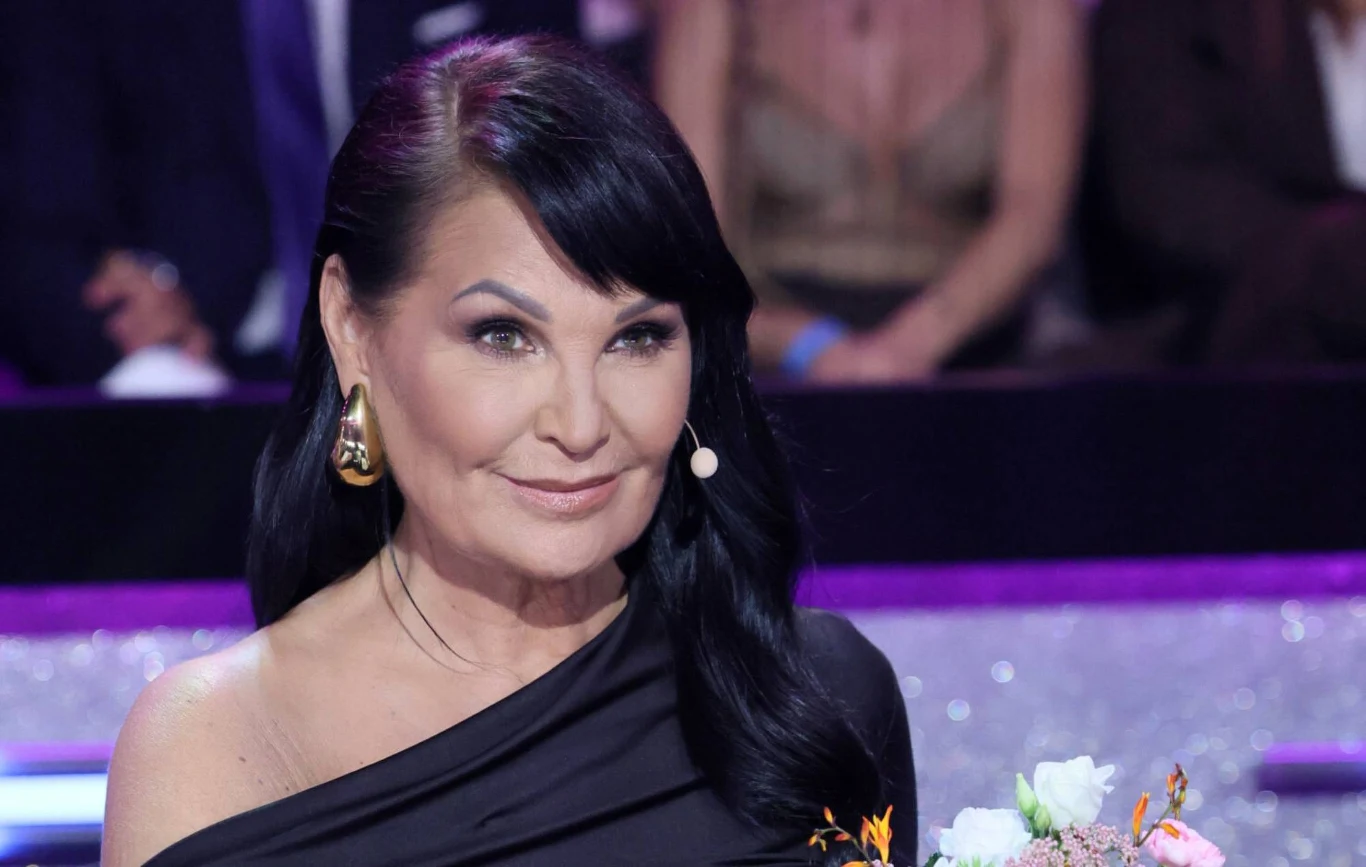 Iwona Pavlović w programie "Taniec z Gwiazdami" Iwona Pavlović w czarnej sukni z odsłoniętym ramieniem, siedzi przy stole w programie 'Taniec z Gwiazdami'.