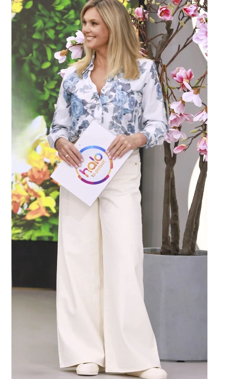 kobieta w jasnych, szerokich spodniach i kwiecistej koszuli trzymająca kolorową teczkę z napisem 'halo', stojąca obok dekoracyjnych gałęzi z różowymi kwiatami