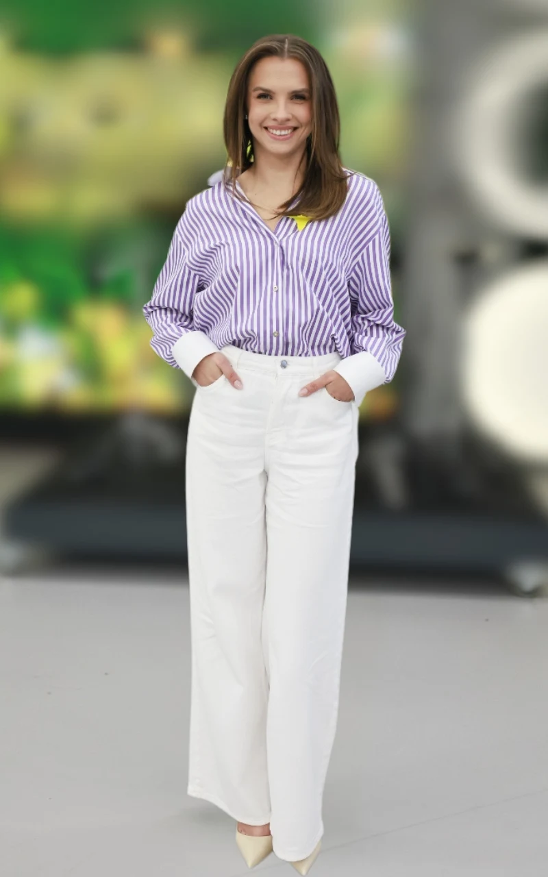 uśmiechnięta kobieta ubrana w koszulę w fioletowe paski i białe szerokie spodnie, stojąca na tle rozmytego zielono-szarego tła