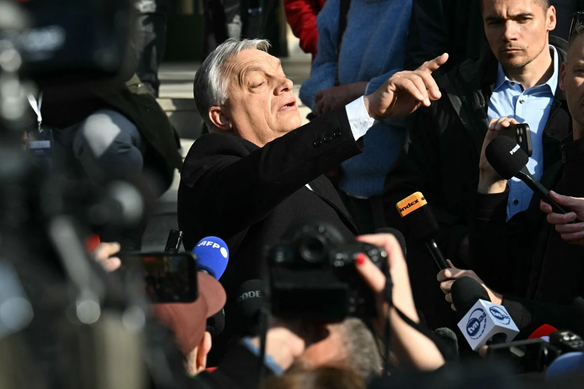 Mężczyzna w garniturze i białej koszuli wskazuje ręką, otoczony przez dziennikarzy z mikrofonami i kamerami, w tłumie ludzi na zewnątrz budynku.