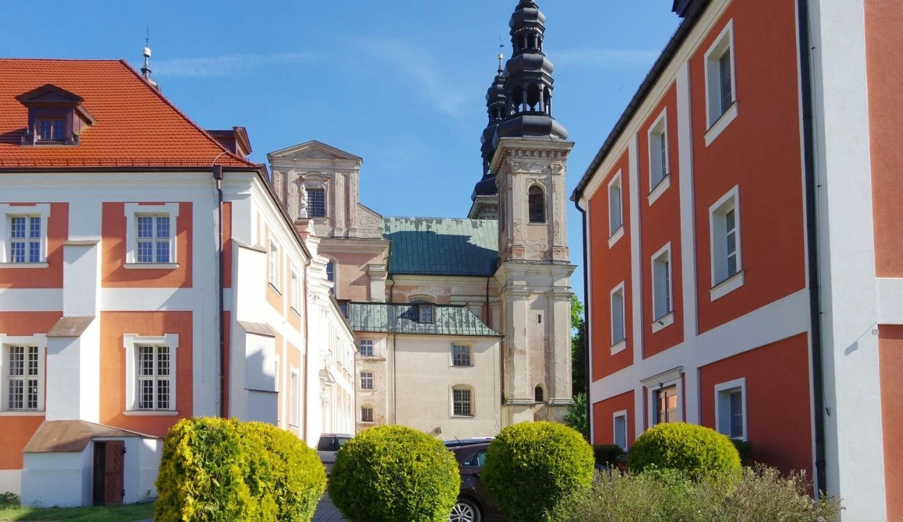 Barokowy kościół z dwoma wysokimi wieżami w tle, otoczony zadbanymi, kolorowymi budynkami o czerwonych i białych elewacjach, na pierwszym planie widoczne gęste krzewy przy jasnozielonym trawniku.