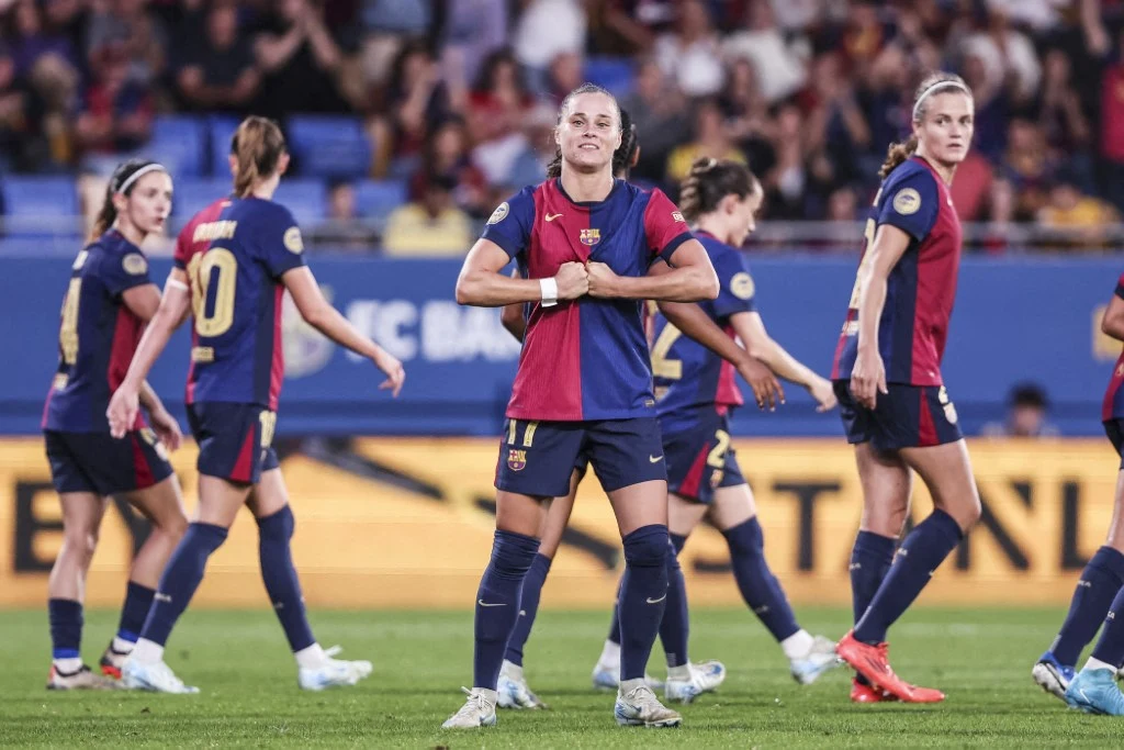 piłkarka w stroju FC Barcelony wykonuje charakterystyczny gest triumfu trzymając koszulkę na klatce piersiowej, otoczona przez koleżanki z drużyny na boisku podczas meczu, w tle widownia na stadionie