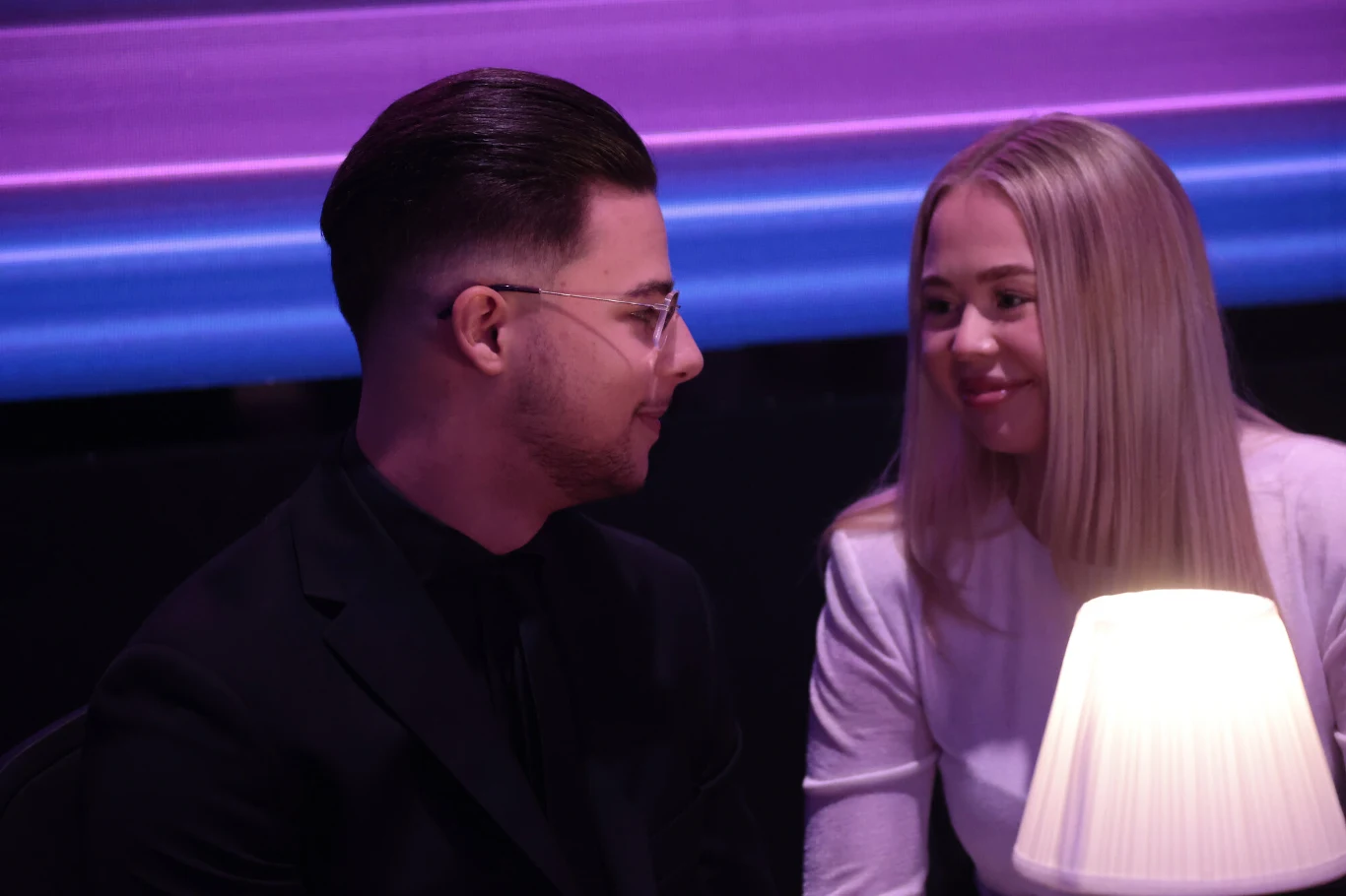 Mężczyzna w okularach i kobieta o blond włosach siedzą razem przy stoliku, patrząc na siebie z uśmiechem, oświetleni ciepłym światłem lampki na tle fioletowych neonów.