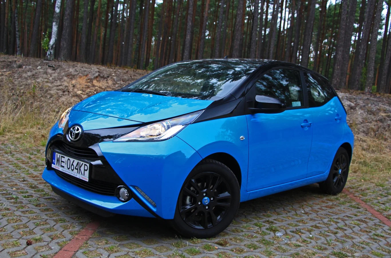 Druga generacja Aygo zadebiutowała w 2014 roku. niebieski samochód Toyota Aygo z czarnymi detalami i felgami stoi zaparkowany na kostce brukowej, w tle widoczny las iglasty