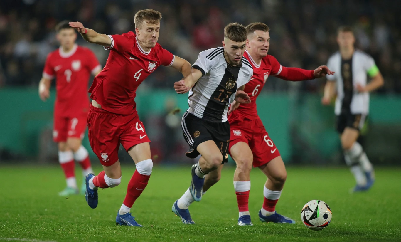 Dwóch piłkarzy w czerwonych strojach próbuje odebrać piłkę zawodnikowi w czarno-białej koszulce podczas dynamicznej akcji na boisku piłkarskim.