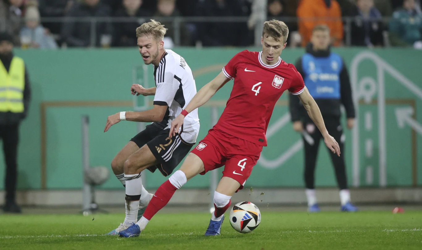 Dwóch piłkarzy rywalizuje o piłkę na boisku, jeden ubrany w biało-czarny strój, drugi w czerwony strój z numerem 4, wokół nich widoczni są inni zawodnicy oraz sędzia na tle trybun i zielonego baneru.