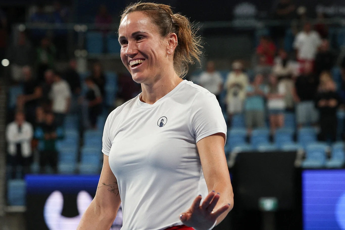 kobieta w białej koszulce sportowej uśmiecha się na korcie tenisowym, w tle rozmyci kibice siedzący na niebieskich trybunach