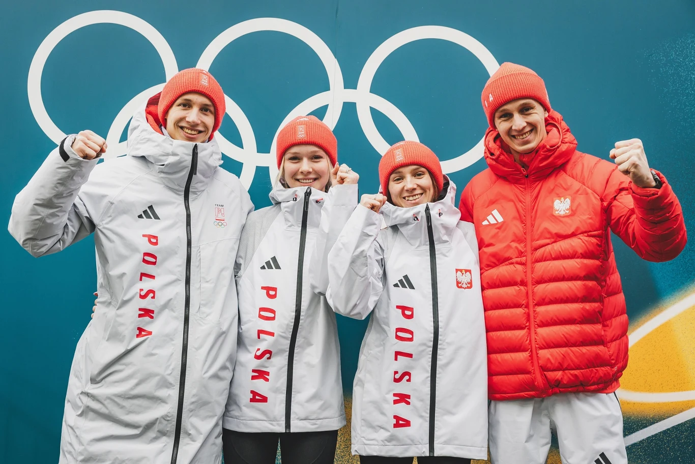 Kacper Tomasiak, Pola Bełtowska, Anna Twardosz, Paweł Wąsek czwórka sportowców ubrana w białe i czerwone kurtki z napisem 'Polska' oraz czapki w barwach narodowych, wszyscy uśmiechają się i trzymają zaciśnięte pięści, w tle widoczne koła olimpijskie