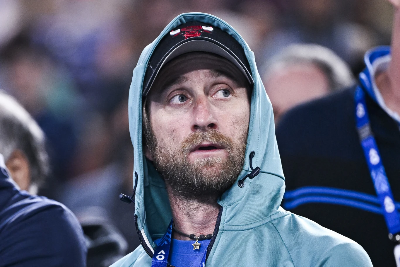 Gilles Cervara Brodaty mężczyzna w sportowej bluzie z kapturem oraz czapce z daszkiem, spoglądający w bok z lekko zaniepokojonym wyrazem twarzy, na tle zamazanego tłumu.