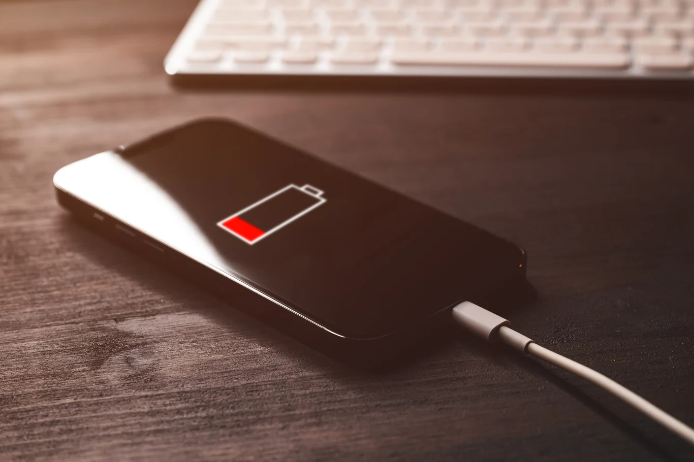 Te nawyki niszczą baterię smartfona smartfon z podłączonym kablem do ładowania, na ekranie symbol niskiego poziomu baterii, w tle klawiatura na ciemnym biurku