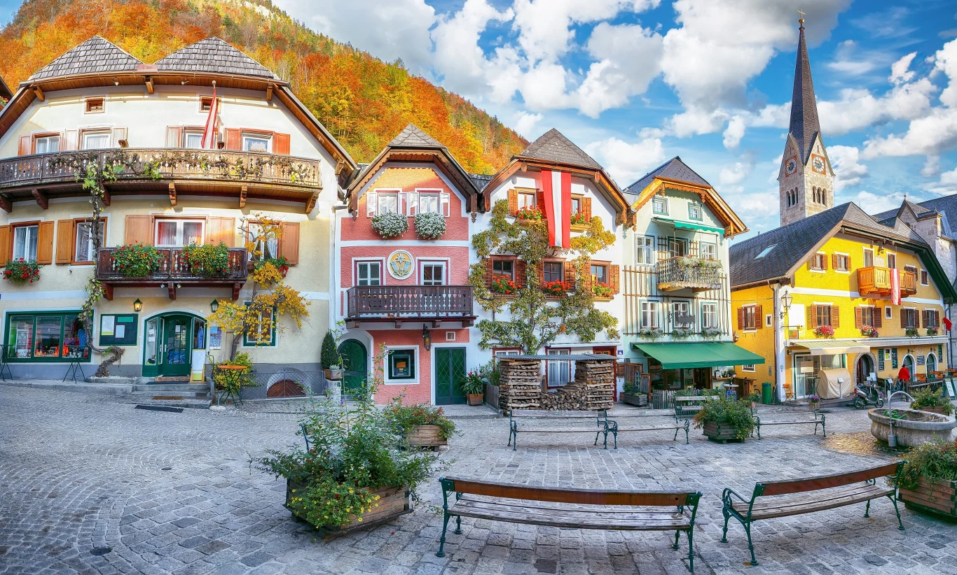 Kolorowe kamienice z drewnianymi balkonami i kwiatami, urokliwy brukowany plac z ławkami i zielenią; w tle góra pokryta jesiennym lasem oraz wieża kościoła z ostrym dachem.