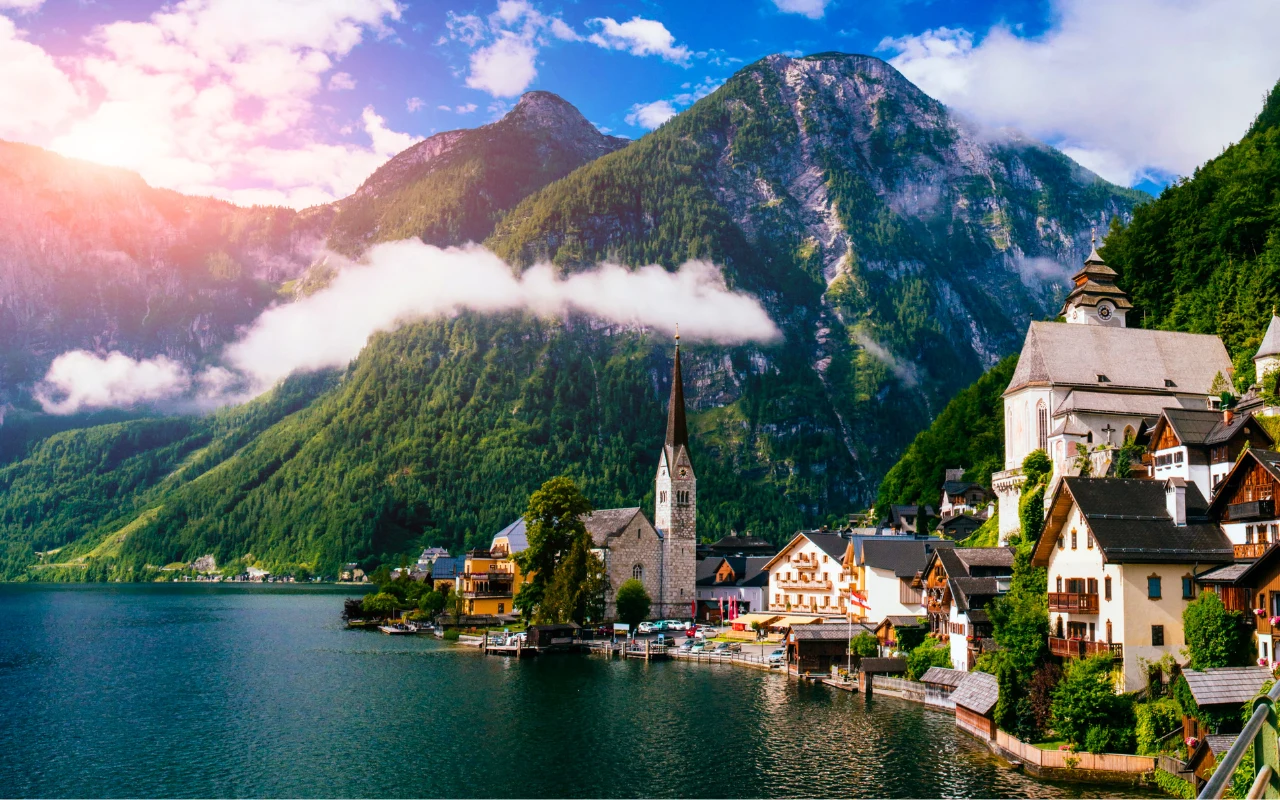 malownicza alpejska miejscowość nad jeziorem, otoczona wysokimi, zielonymi górami, z charakterystycznym kościołem o wysokiej wieży i tradycyjną zabudową położoną blisko brzegu
