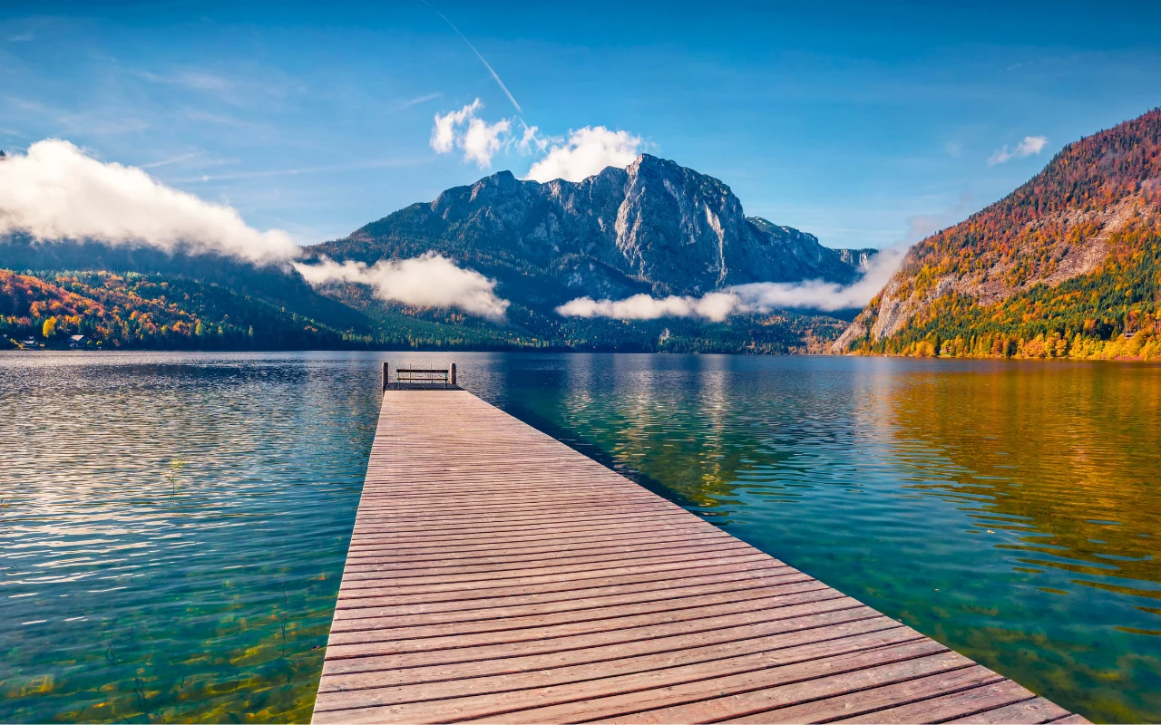 drewniane molo prowadzi wprost na spokojną taflę jeziora, otoczonego górami porośniętymi jesiennym lasem, a nad wodą unoszą się niskie chmury, podkreślając majestatyczną sylwetkę skalistego szczytu w tle