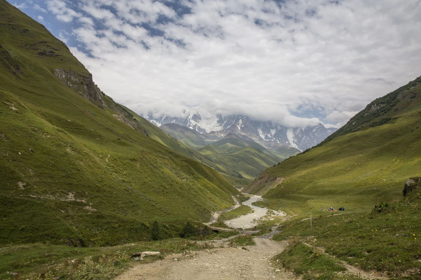 kręta, kamienista ścieżka biegnąca przez zieloną dolinę otoczoną wysokimi wzgórzami, w oddali ośnieżone szczyty gór częściowo zasłonięte chmurami
