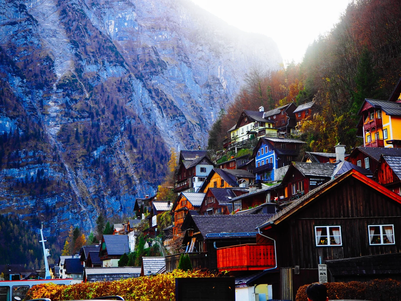 Kolorowe, drewniane domy o stromych dachach ułożone tarasowo na zboczu góry, otoczone jesienną roślinnością, w tle imponujący skalisty masyw górski podświetlony światłem dziennym.