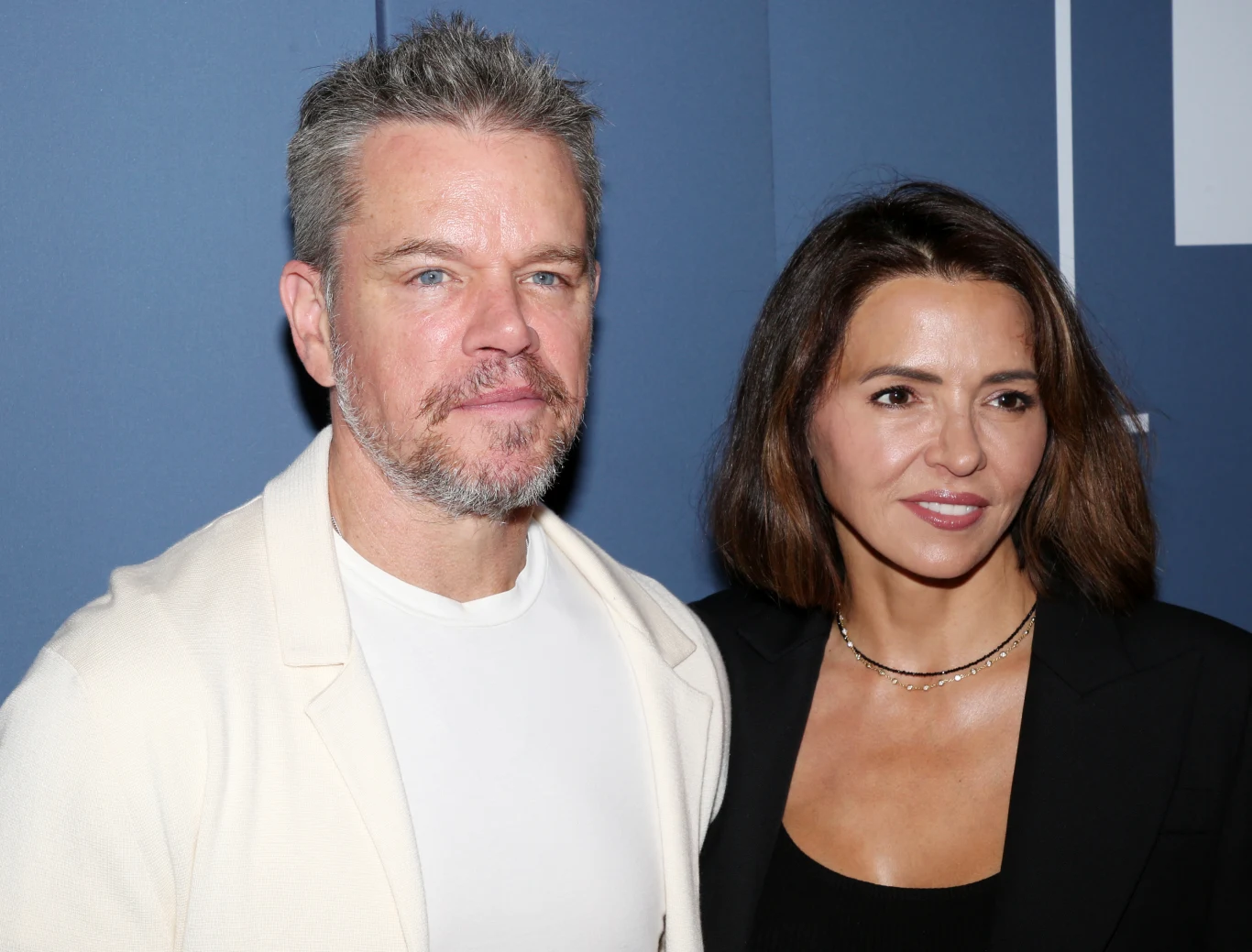 Matt Damon i Luciana Barroso Damon Mężczyzna z krótkimi włosami i zarostem w swetrze oraz kobieta o ciemnych włosach stoją razem na tle niebieskiej ściany
