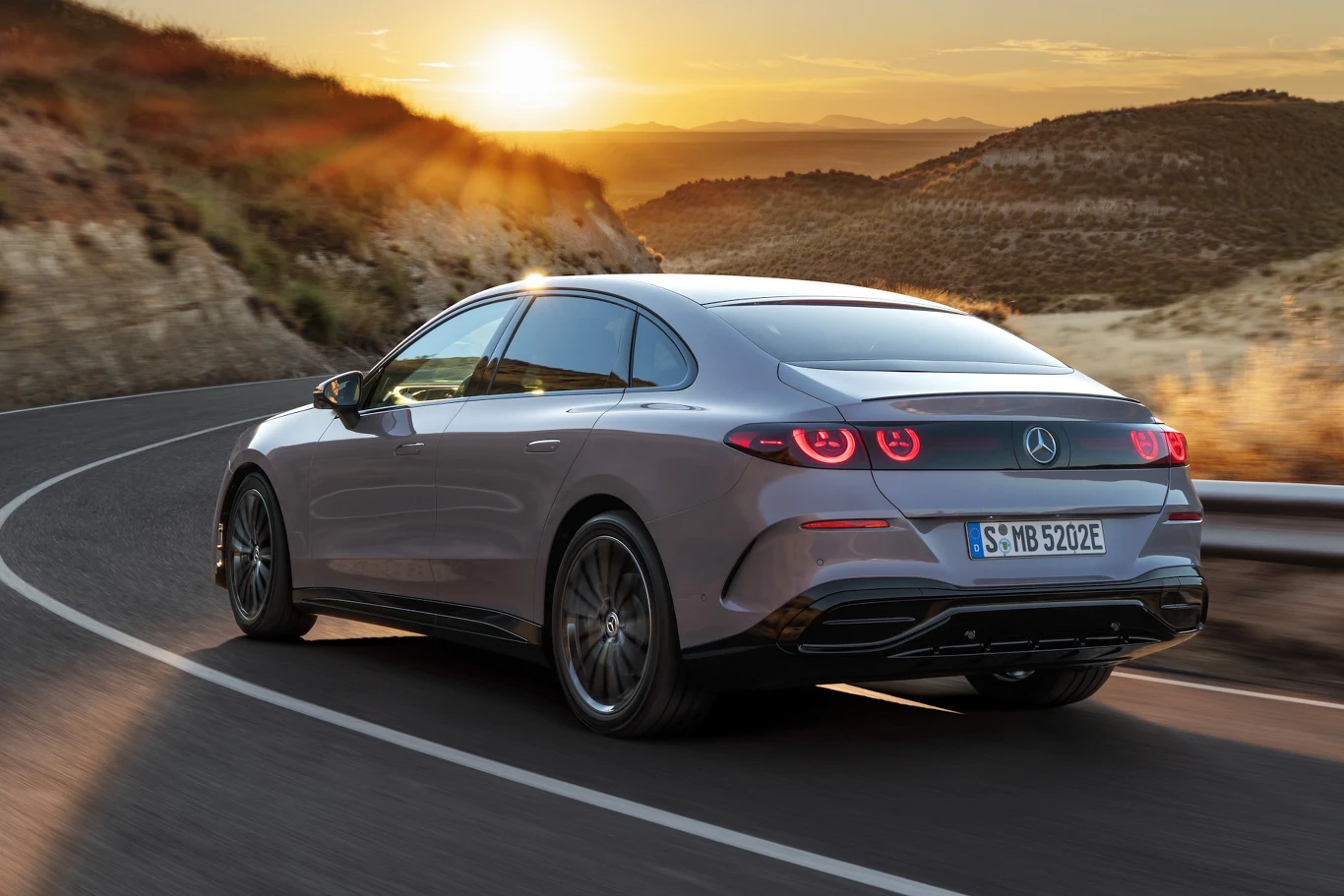 Mercedes klasy C Electric Nowoczesny szary samochód marki Mercedes poruszający się krętą, asfaltową drogą wśród górskiego krajobrazu o zachodzie słońca, z widocznymi czerwonymi światłami tylnymi i aerodynamiczną sylwetką.