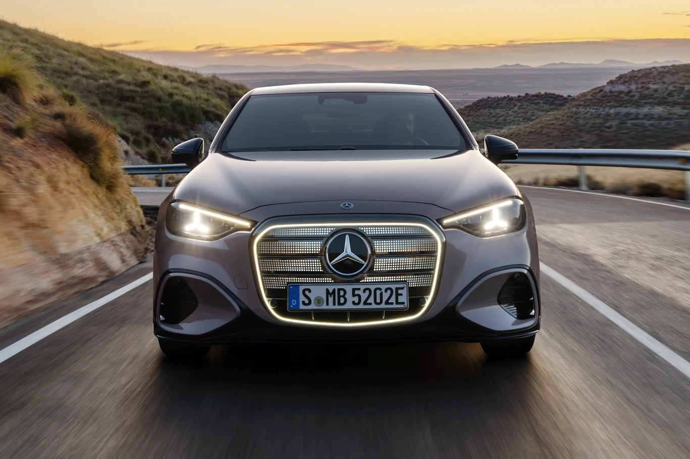 Mercedes klasy C Electric luksusowy samochód marki Mercedes z charakterystycznym podświetlanym grillem i nowoczesnymi reflektorami, jadący pustą, krętą drogą w górskim krajobrazie o zachodzie słońca