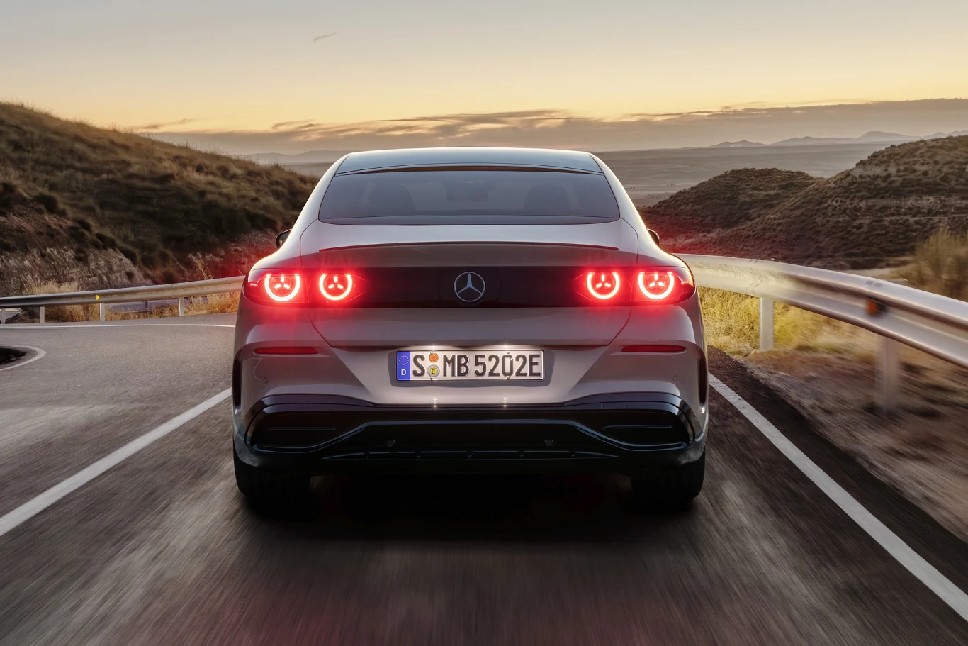 Mercedes klasy C Electric nowoczesny samochód marki Mercedes z włączonymi światłami tylnymi poruszający się po krętej, asfaltowej drodze otoczonej pagórkowatym krajobrazem o zachodzie słońca