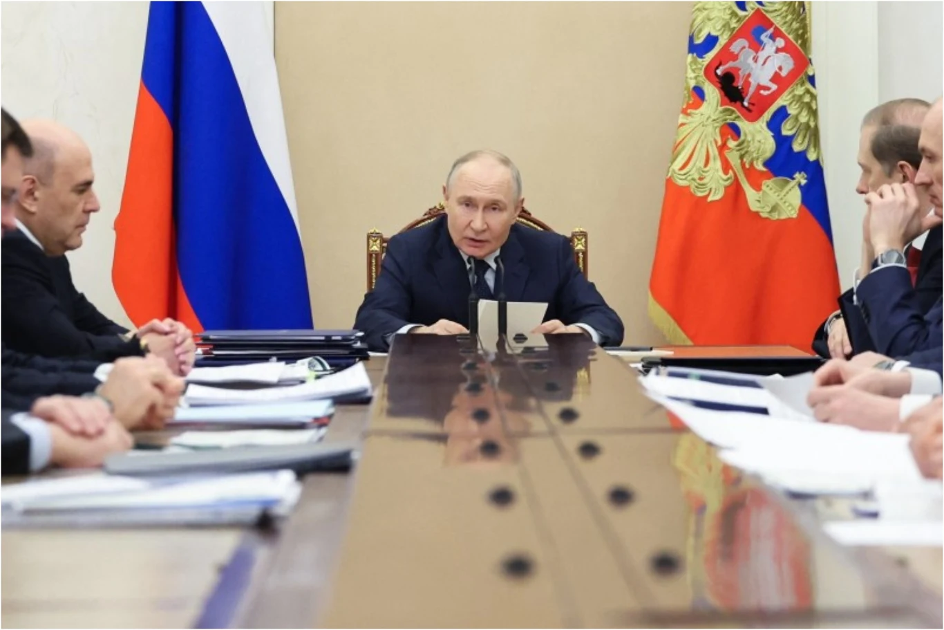 Prezydent Rosji Władimir Putin przewodniczył spotkaniu poświęconemu kwestiom gospodarczym Władimir Putin na spotkaniu gospodarczym, siedzi przy długim stole, w tle flaga Rosji i herb, na stole dokumenty.