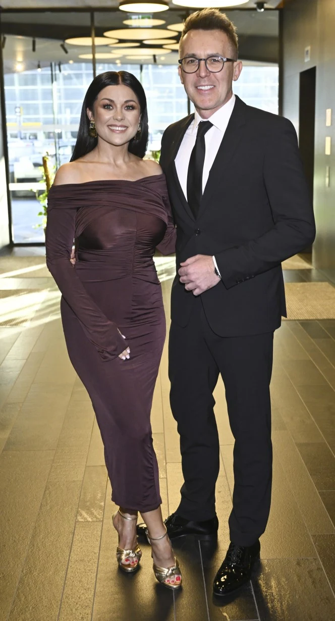 Elegancko ubrana kobieta w długiej, bordowej sukni wieczorowej i złotych sandałkach stoi obok mężczyzny w czarnym garniturze, białej koszuli i krawacie; oboje uśmiechnięci pozują w nowoczesnym, dobrze oświetlonym wnętrzu.