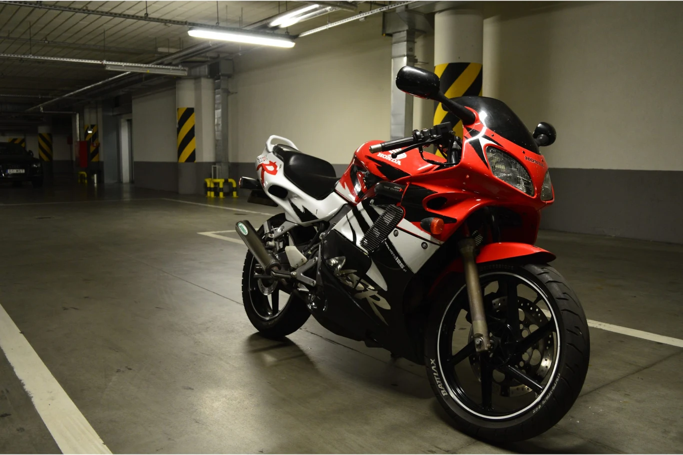 Jakie są najszybsze motocykle 125? Wśród najszybszej trójki znajduje się Honda NSR 125. Honda NSR 125 w czerwono-białych barwach zaparkowana w oświetlonym garażu podziemnym z żółto-czarnymi kolumnami