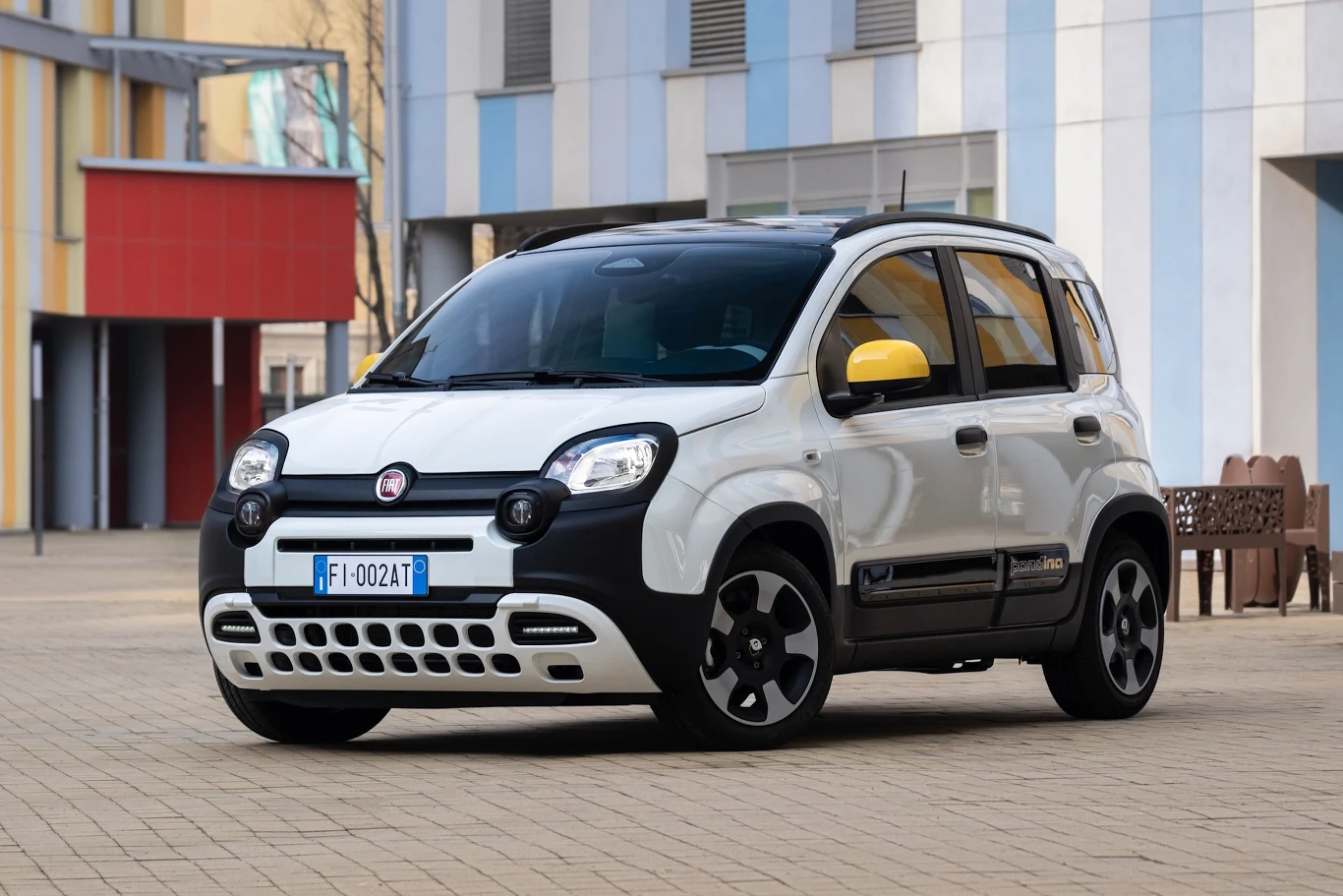 biały Fiat Panda z czarnymi i żółtymi akcentami parkuje na brukowanym placu przy nowoczesnym budynku o niebiesko-białych elewacjach