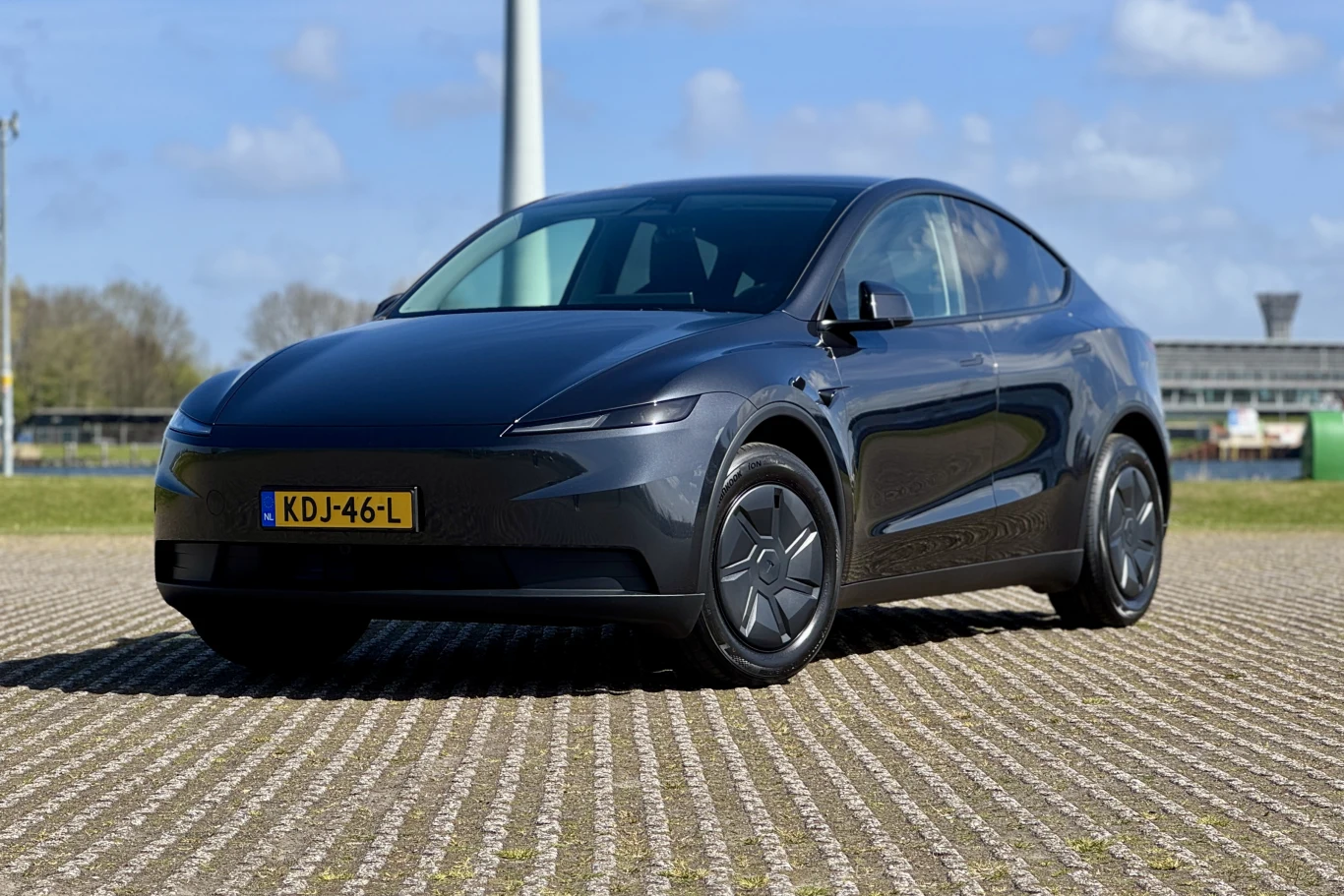 Czarny nowoczesny samochód elektryczny o aerodynamicznym kształcie stoi na parkingu, z żółtymi tablicami rejestracyjnymi i gładkimi felgami, w tle widać fragment niskich budynków i niebo z chmurami.