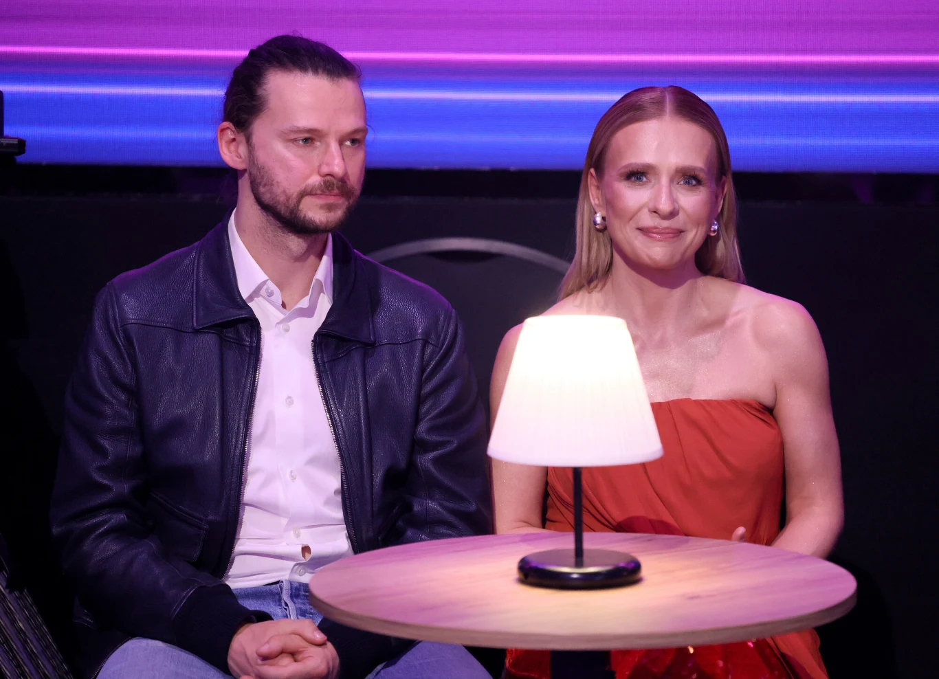 dwoje ludzi siedzi przy okrągłym stoliku oświetlonym lampką, mężczyzna w skórzanej kurtce i kobieta w czerwonej sukience, w tle fioletowe światła