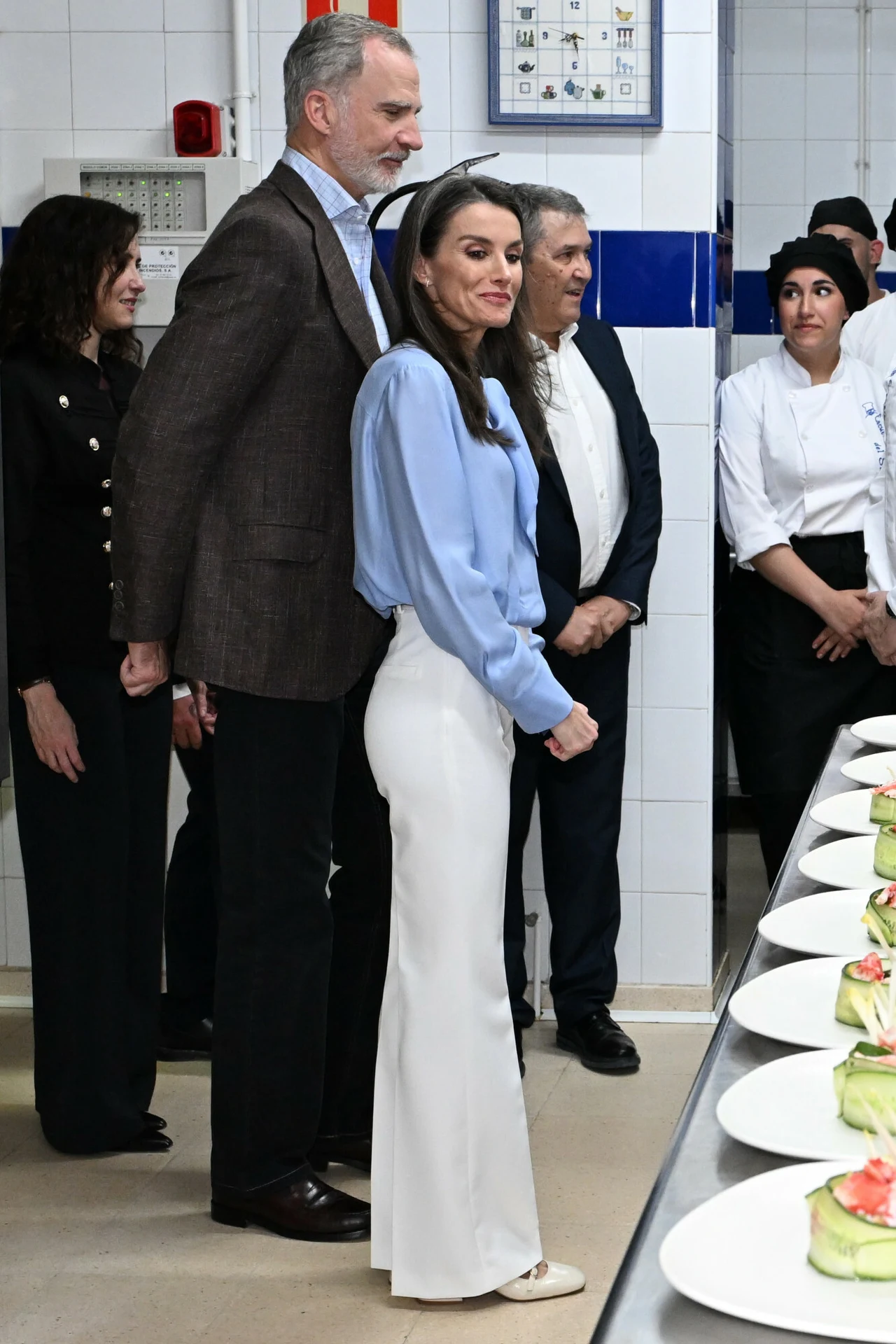 Para elegancko ubranych osób stoi w kuchni obok stołu, na którym ułożono talerze z przystawkami; w tle kilka osób w garniturach i kucharskich uniformach obserwuje sytuację.