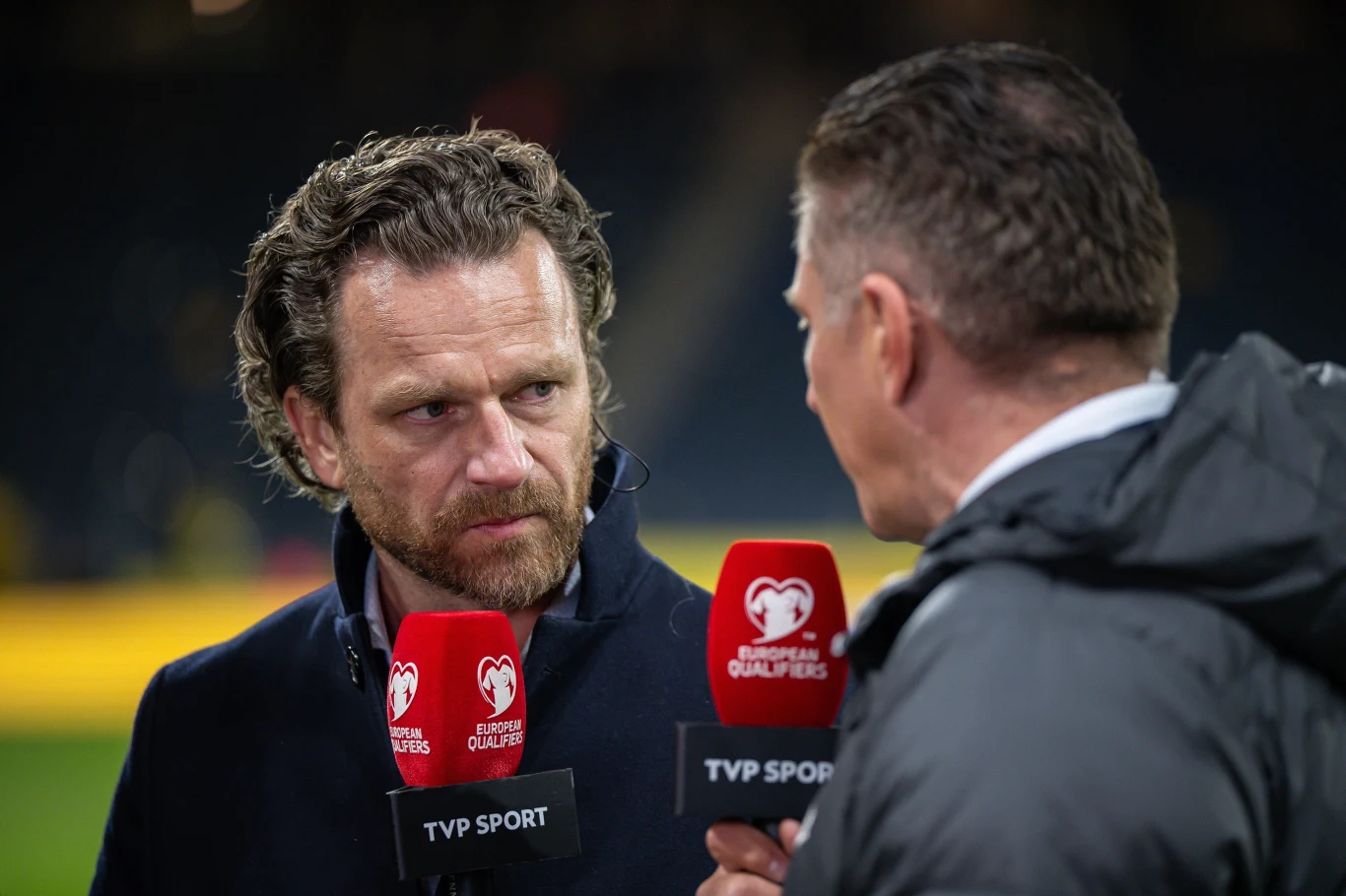 Dwóch mężczyzn prowadzi wywiad na tle stadionu, trzymając czerwone mikrofony oznaczone logo European Qualifiers oraz TVP Sport; po lewej stronie postać o poważnym wyrazie twarzy, po prawej drugi mężczyzna z profilu.