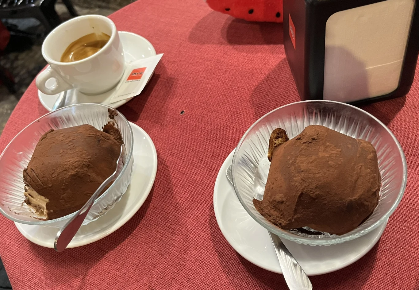 Będąc w Tropei albo w Pizzo koniecznie trzeba spróbować słynnego lodowego deseru - tartuffo Dwie szklane miseczki z porcjami tiramisu posypanymi kakao oraz filiżanka espresso na czerwonym obrusie, obok serwetnik.