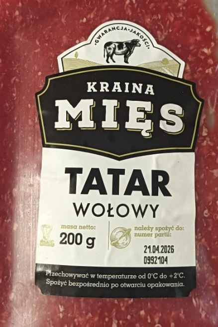 opakowanie wołowego tatara marki Kraina Mięs o wadze 200 g z wyraźnie widoczną etykietą informującą o dacie przydatności i zaleceniach przechowywania, na tle surowego mięsa