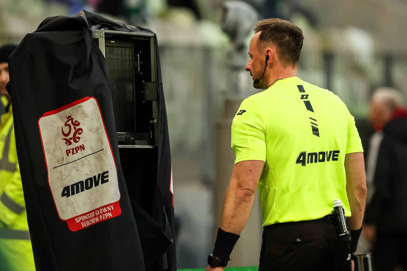 sędzia piłkarski w żółtym stroju stoi obok monitora VAR osłoniętego czarnym pokrowcem z logotypem PZPN i napisem 'Sędzia Główny Sędziuje Zewn'